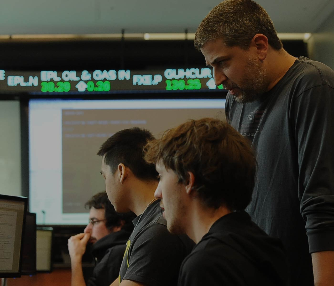 Professor advising students with stock exchange reading in background