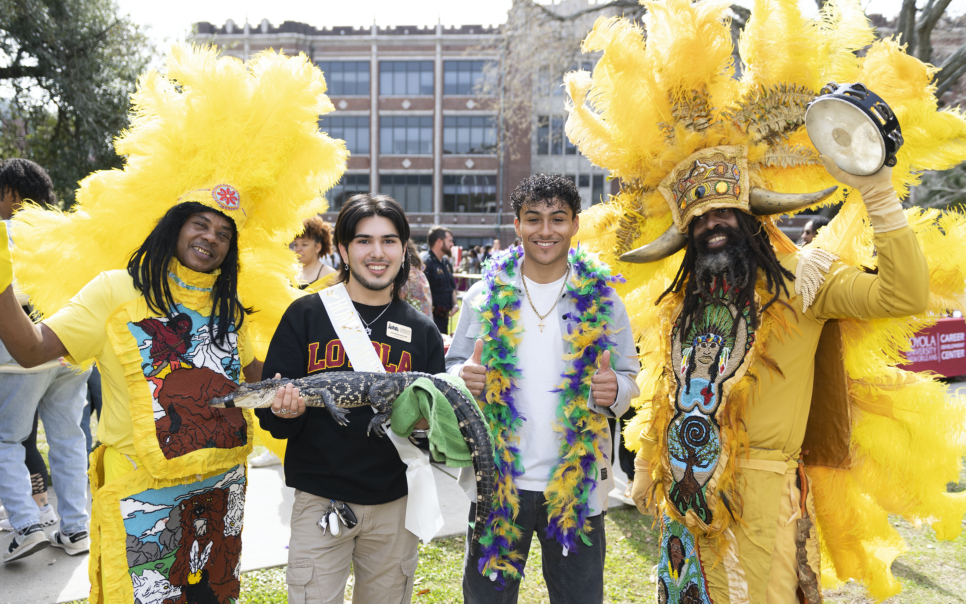 Two students, one holding a baby alligator and another with a feather boa on posing with Mardi Gras Indians.
